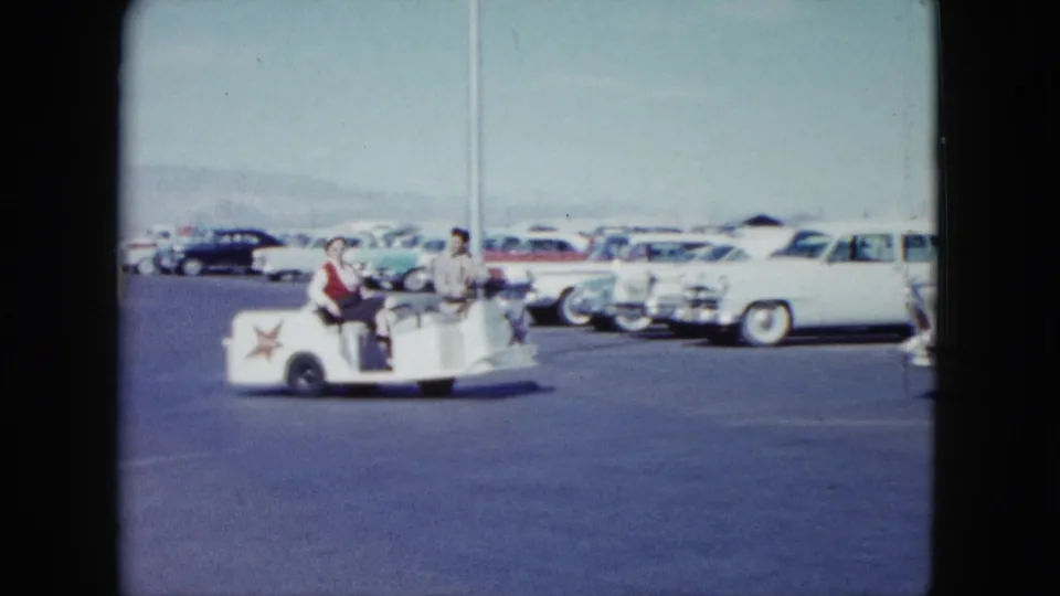 The white guide cart rolls past a large white 1950s car in Nevada, with parked vehicles and mountains across the background.
