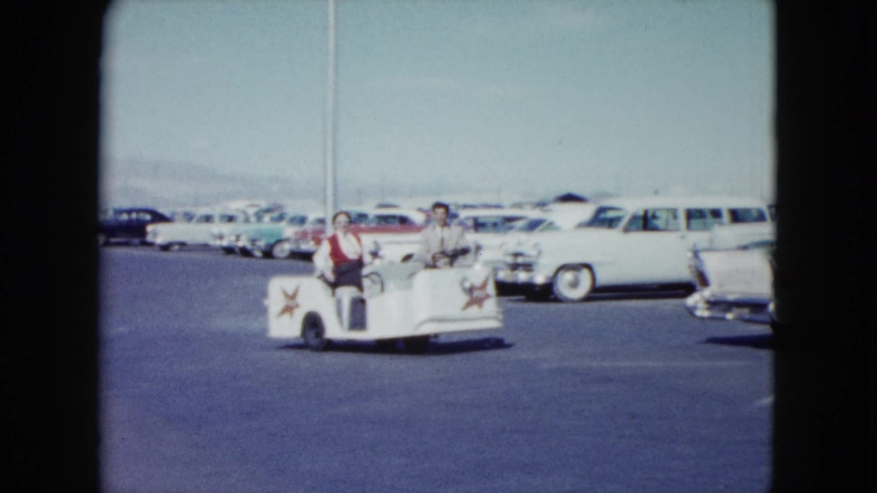 Two adults ride a small white utility cart through a parking lot lined with classic cars.