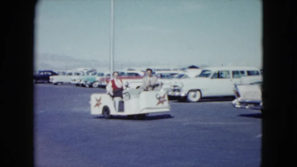In 1959 Nevada home-movie footage, a man and woman ride a white utility cart past rows of late-1950s cars in a sunlit parking lot.