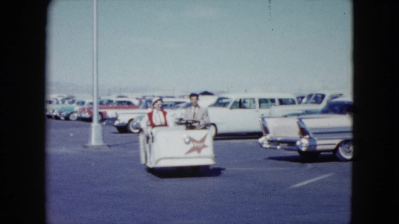 A man drives a white cart with a woman beside him, passing parked cars and a light pole.