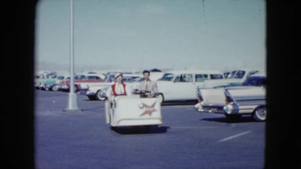 The couple rides closer in the white utility cart, with red star markings visible and late-1950s cars parked across the Nevada lot.