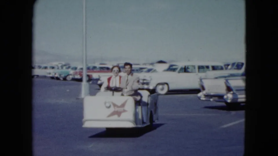 A man and woman in period clothing ride a small white cart through a Nevada parking lot, framed by light poles and parked 1950s cars.