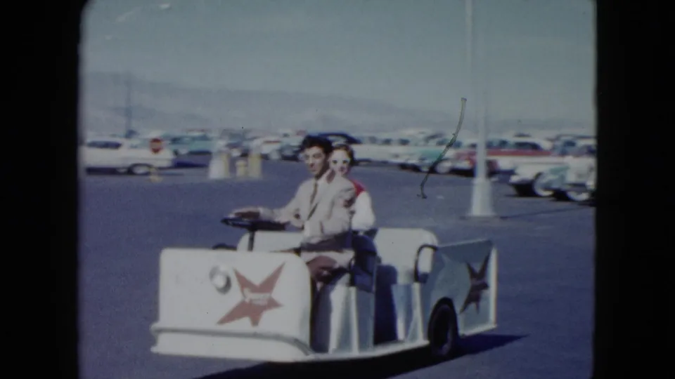 The cart passes nearer the camera, showing the driver, passenger, red star markings, and a broad parking lot of classic cars in 1959 Nevada.
