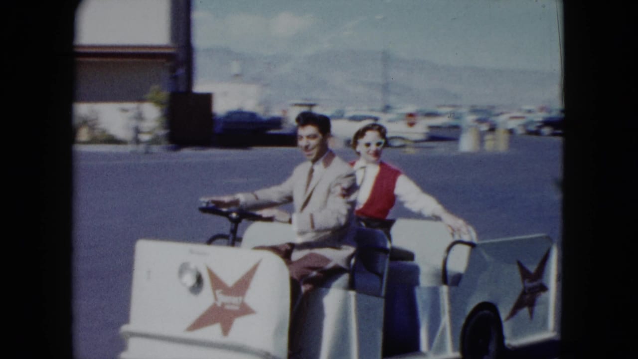A smiling couple rides in a white golf cart with red star logos in a sunny parking lot.
