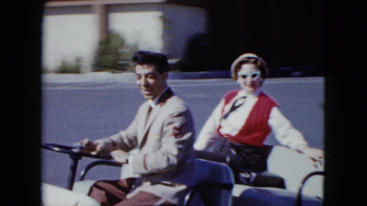 The couple passes close to camera in the white cart, with the woman's sunglasses and red vest clearly visible.