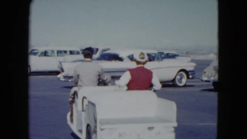 From behind, the couple rides the white golf cart across a Nevada parking lot toward rows of classic cars.