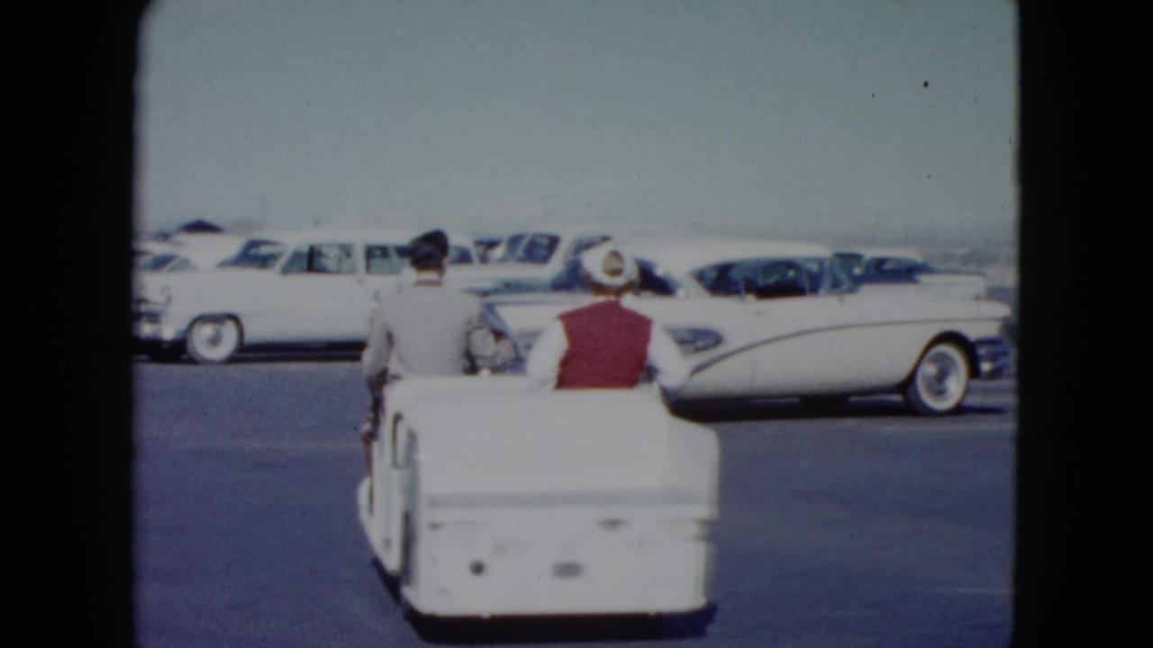 In 1959 Nevada footage, a woman in a red vest and white hat rides a small white utility cart with a suited man past parked classic cars.