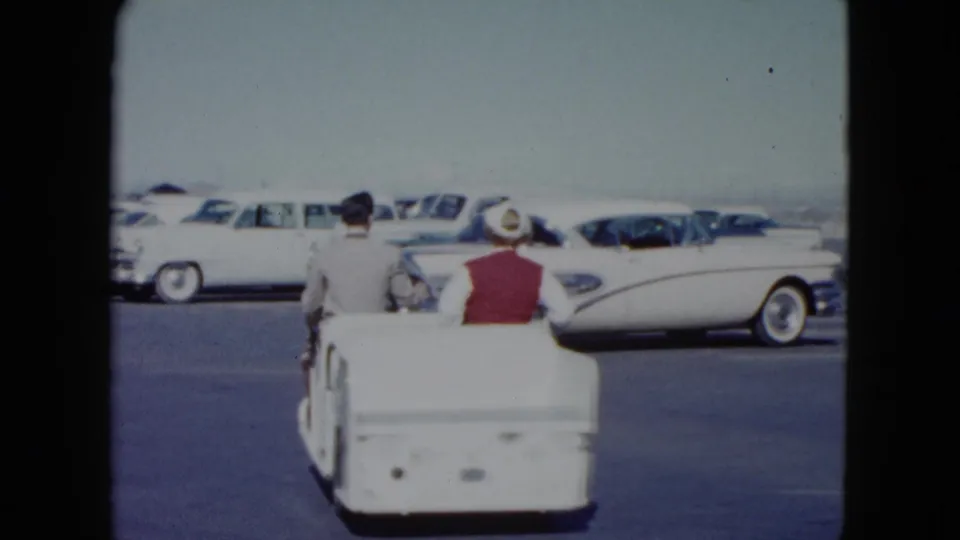 In 1959 Nevada footage, a woman in a red vest and white hat rides a small white utility cart with a suited man past parked classic cars.
