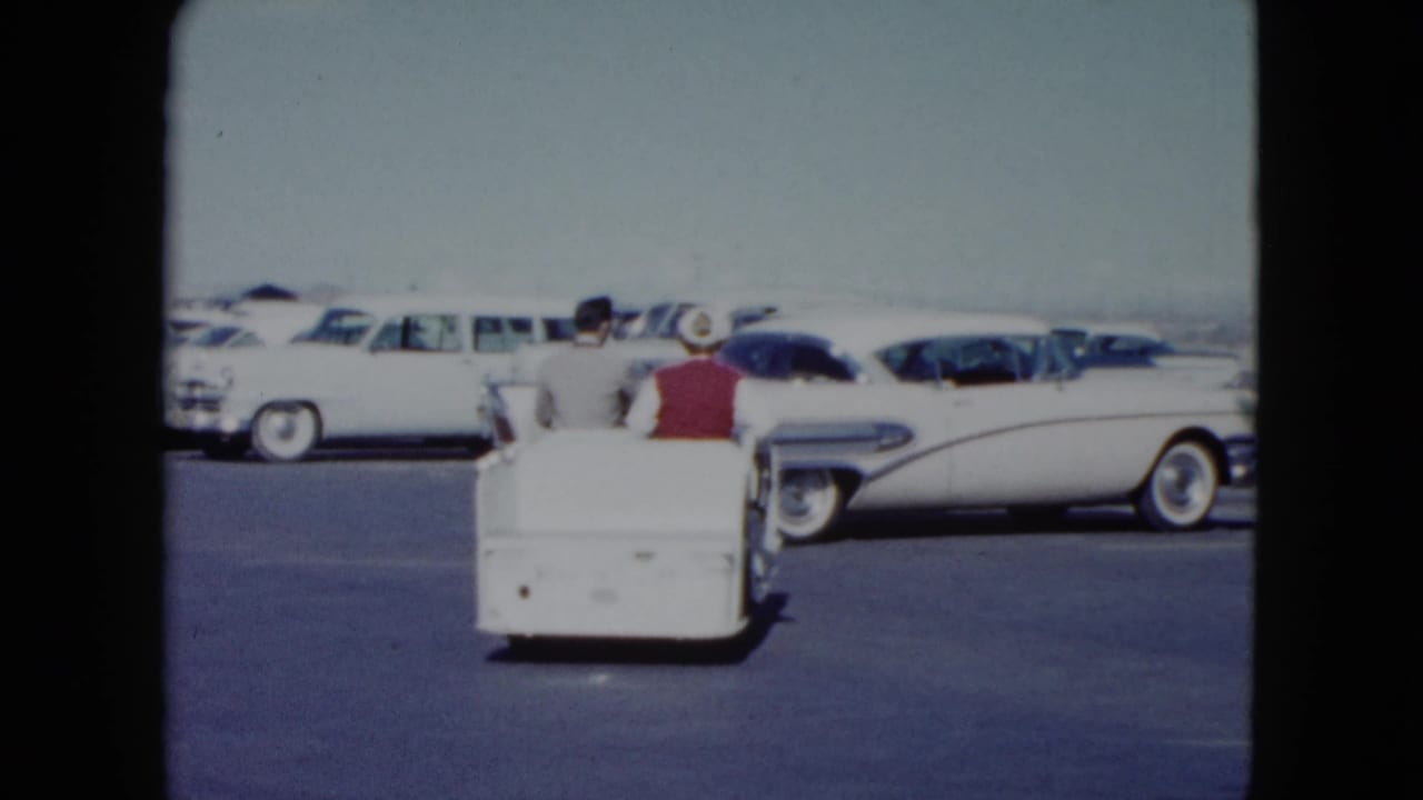The small utility cart continues across the Nevada parking lot, with two adults seated and rows of late-1950s cars behind them.