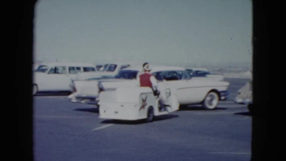 The white utility cart turns through the 1959 Nevada parking lot, carrying the woman in red past a two-tone car and other parked vehicles.