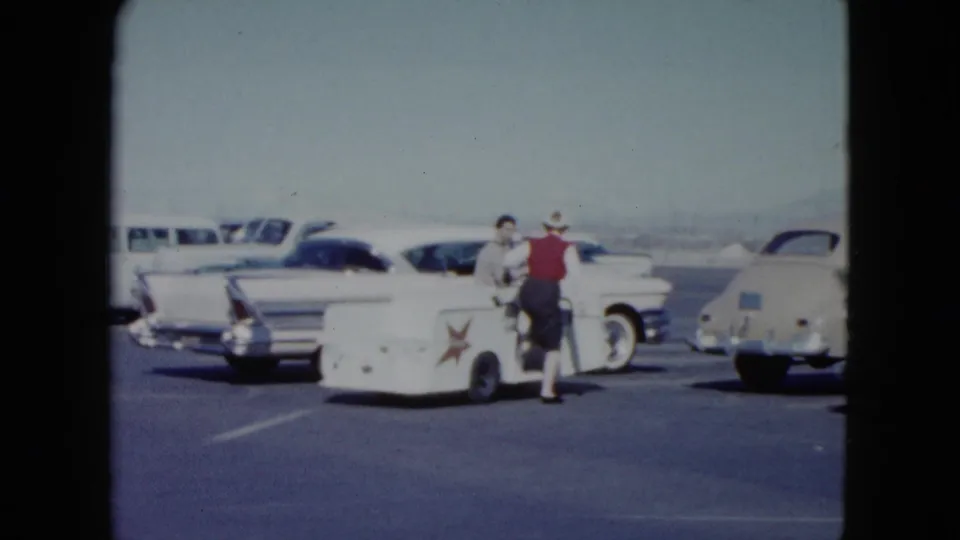 At the end of the clip, the woman in the red vest stands beside the small cart with a suited man amid classic cars in the Nevada lot.
