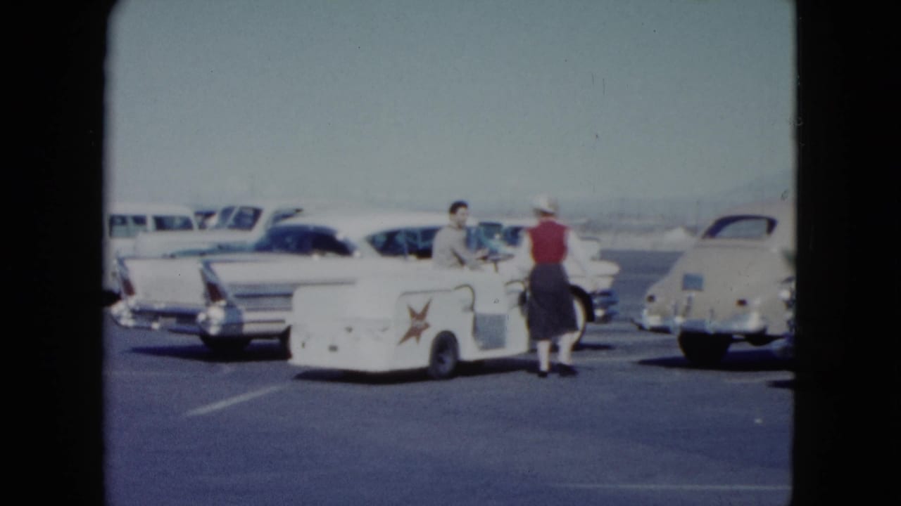 Woman in red vest approaches a man with a white star-marked luggage cart among parked 1950s cars.