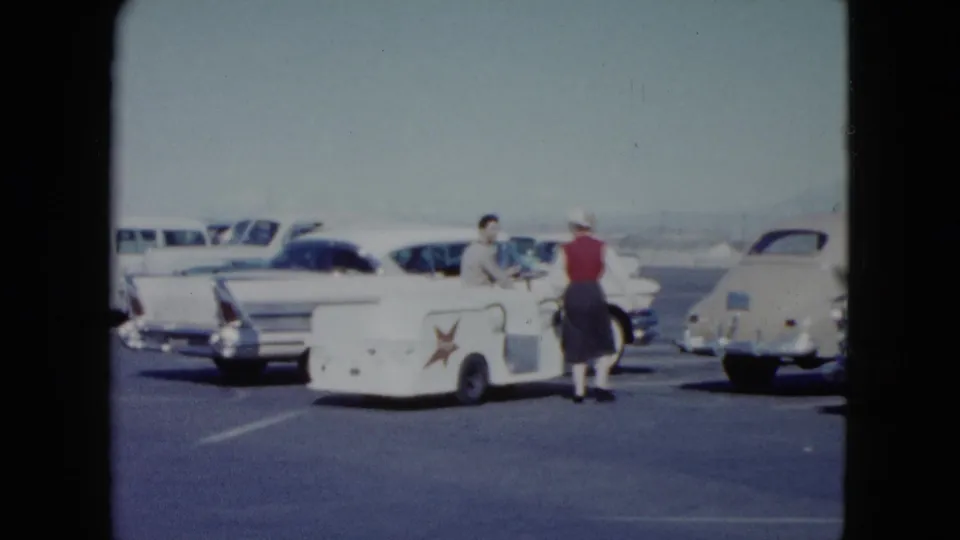At a Nevada airport parking lot in 1959, a woman in a red vest and white hat walks toward a man beside a white service cart.