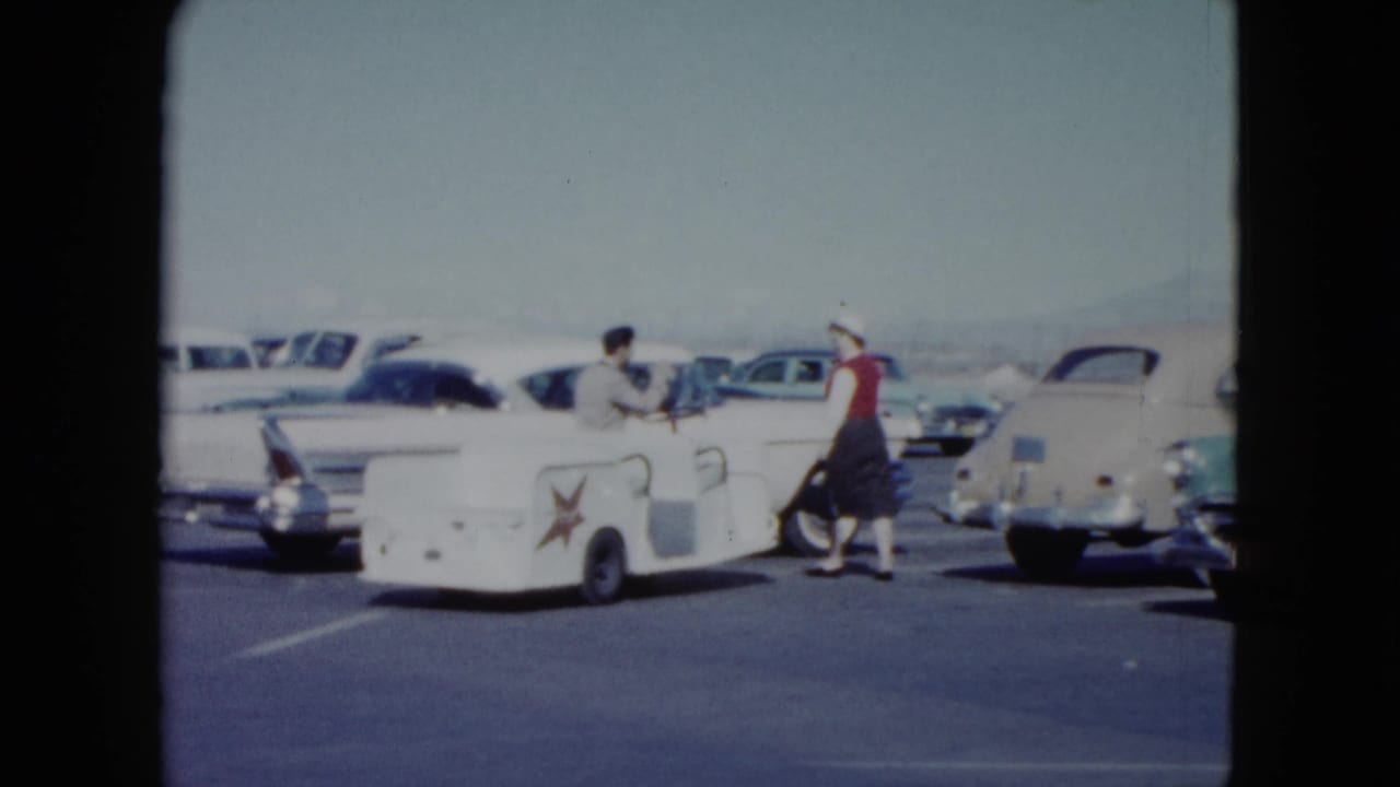 The woman moves through the Nevada airport lot toward a white tailfin sedan while a man remains by the service cart.