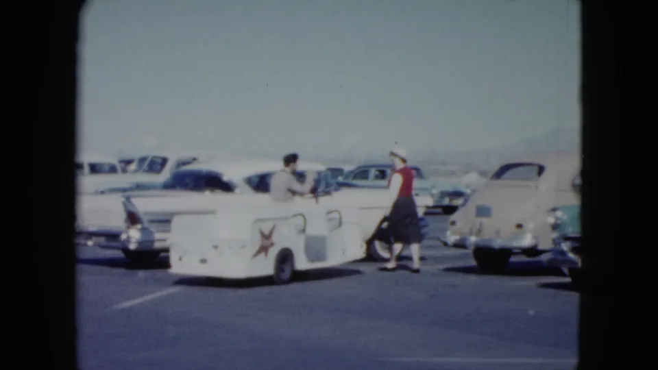 The woman moves through the Nevada airport lot toward a white tailfin sedan while a man remains by the service cart.