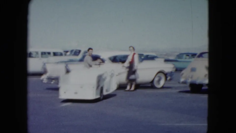 A man moves the white star-marked cart as the woman stands beside a classic white car in the 1959 Nevada airport lot.