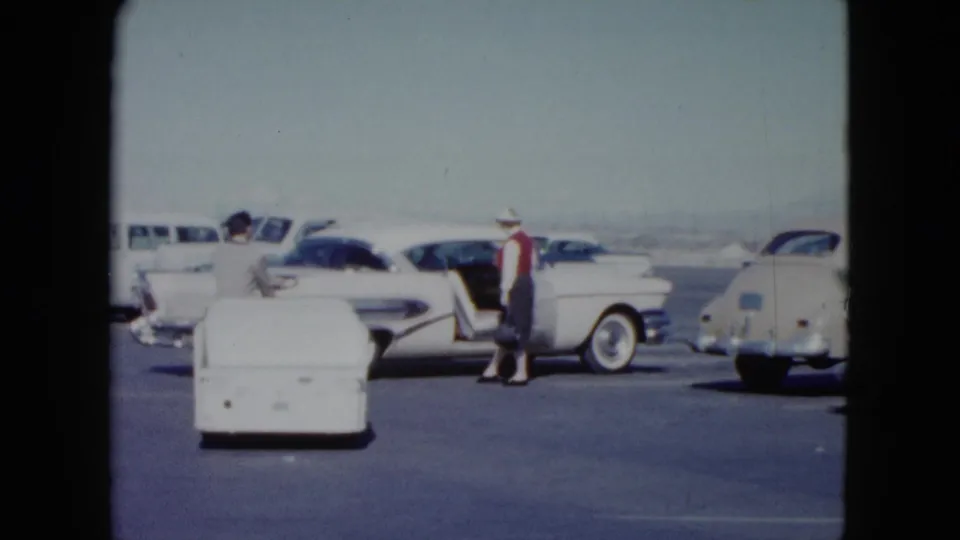 The woman waves near a white tailfin sedan as the man with the small cart turns away in the Nevada airport parking lot.
