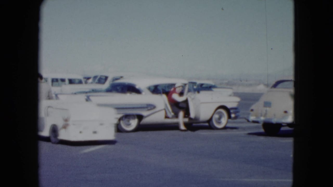 1959 Nevada frame with a woman at the open door of a white classic sedan in a crowded roadside parking area.