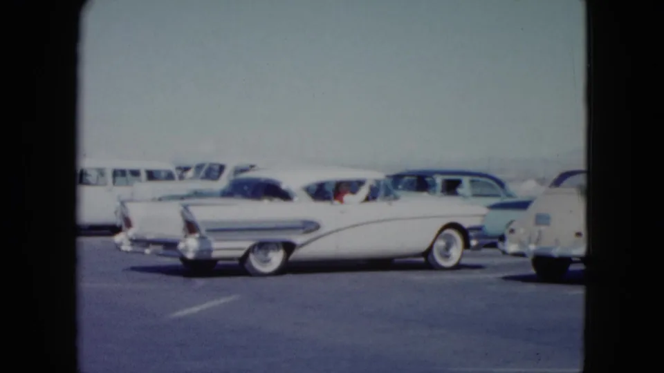 1959 Nevada frame showing a white 1950s sedan beside a pale blue car and other parked vehicles at a roadside stop.