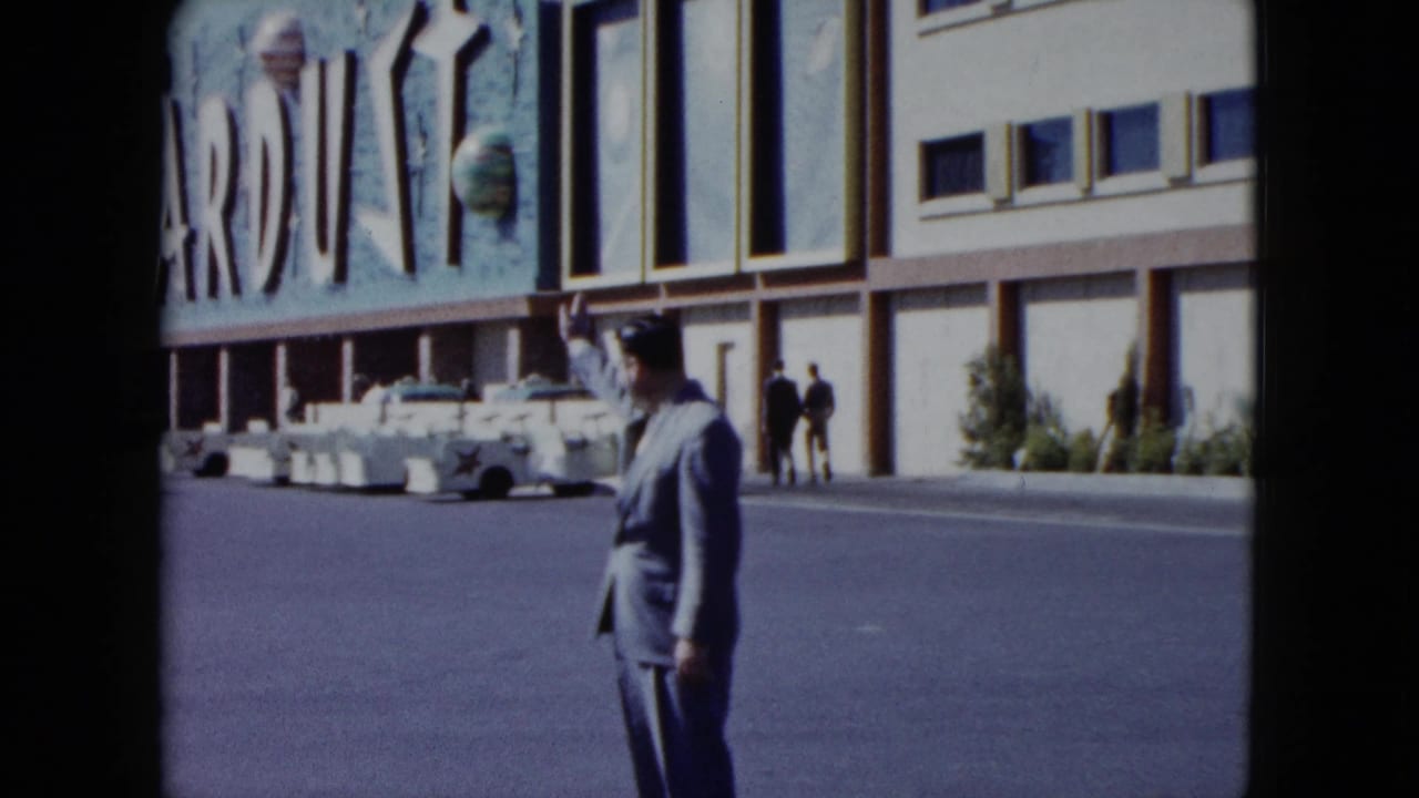 Man in a light suit waves near a street in front of a building with a partial sign.