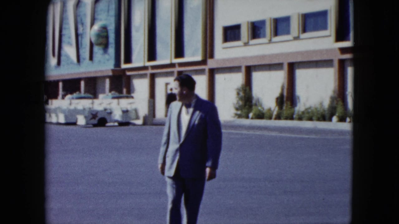 Man in a light suit stands on the street with parked cars and a large sign behind him.