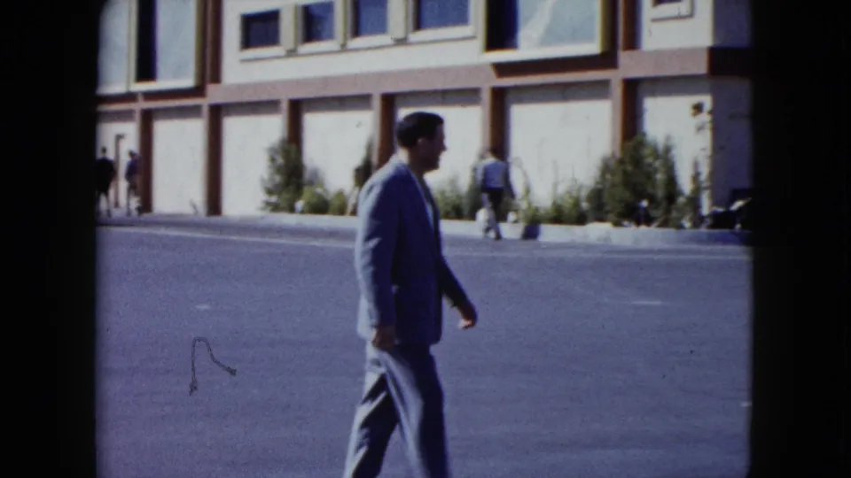 The suited man walks left to right along the Nevada street, with pedestrians and the building facade visible in the background.