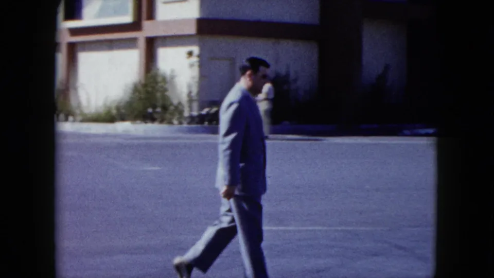 The man continues walking across the frame on a sunlit Nevada street, with the building frontage and roadside planting behind him.