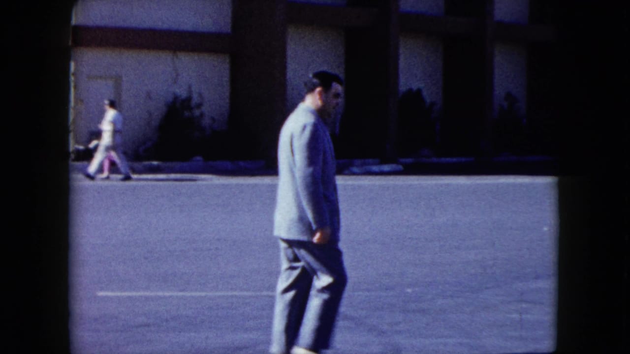 In Nevada home-movie footage from 1959, a suited man crosses a paved lot with a building facade and distant pedestrian behind him.