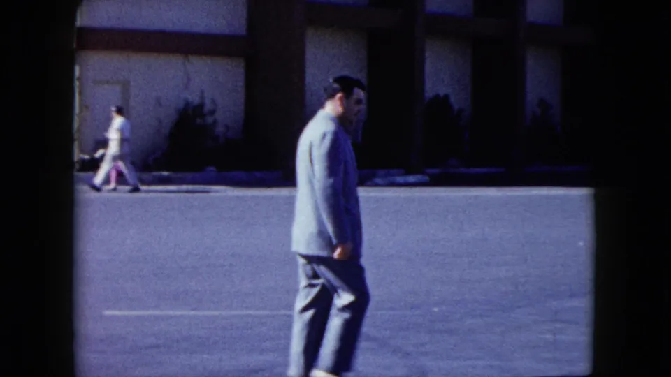 In Nevada home-movie footage from 1959, a suited man crosses a paved lot with a building facade and distant pedestrian behind him.