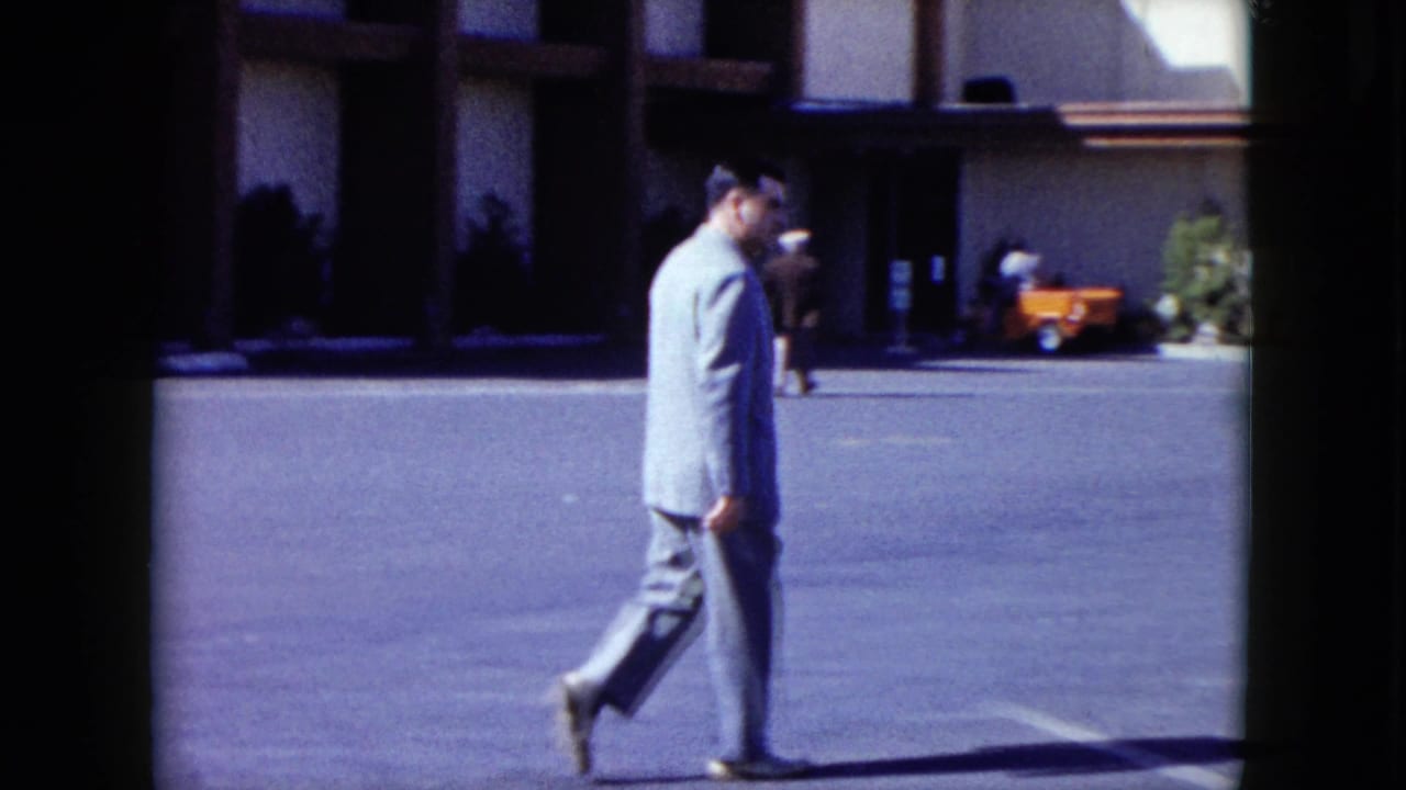 Suited man continues walking across the lot with pedestrians and an orange cart in the background.