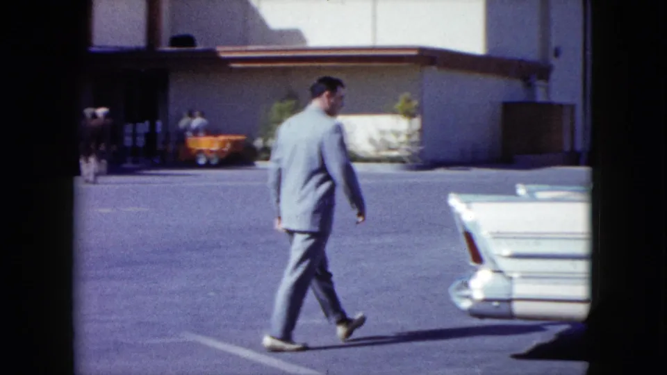 The 1959 Nevada clip shows the suited man nearing a white mid-century car with prominent rear fins, with the building and orange cart in the background.