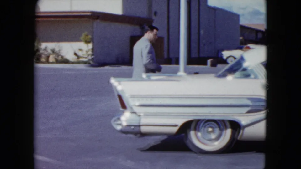 A white finned automobile dominates the foreground while the suited man passes behind it in the Nevada parking lot scene.