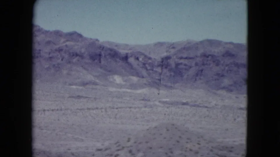 1959 Nevada 8mm frame showing arid hills, sparse vegetation, dry ground, and a visible vertical film scratch or mark.