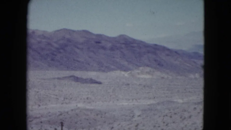1959 Nevada 8mm travel frame with a low desert mound, scrubby foreground, and a continuous mountain range in the distance.