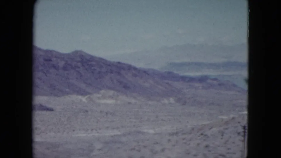 1959 Nevada 8mm frame showing the pan reaching hazier desert ridges, with scrubland foreground and faded blue-purple color.