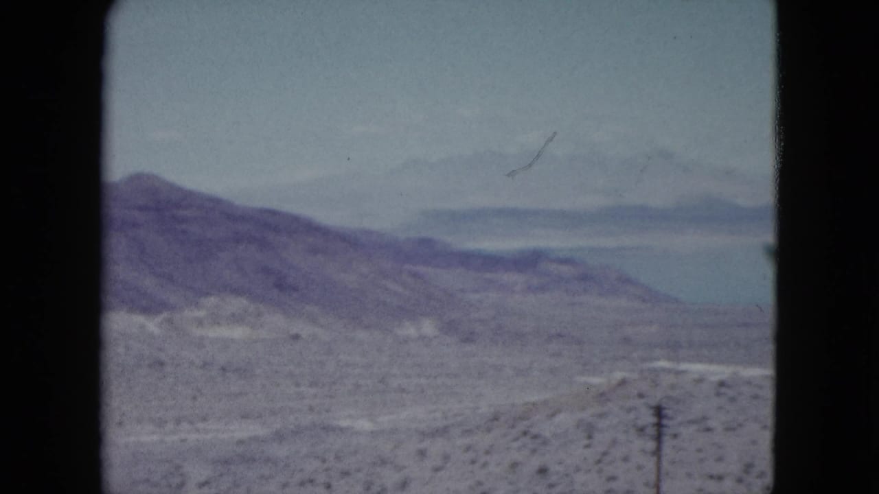 1959 Nevada 8mm frame showing arid hills, a pale lake, distant mountains, scrubland, and a utility pole in the foreground.