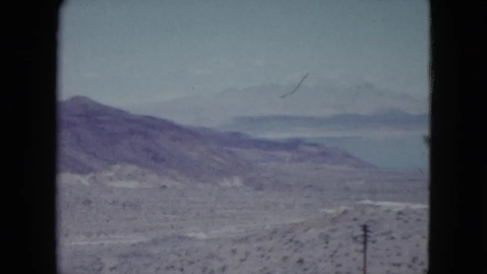1959 Nevada 8mm frame showing arid hills, a pale lake, distant mountains, scrubland, and a utility pole in the foreground.