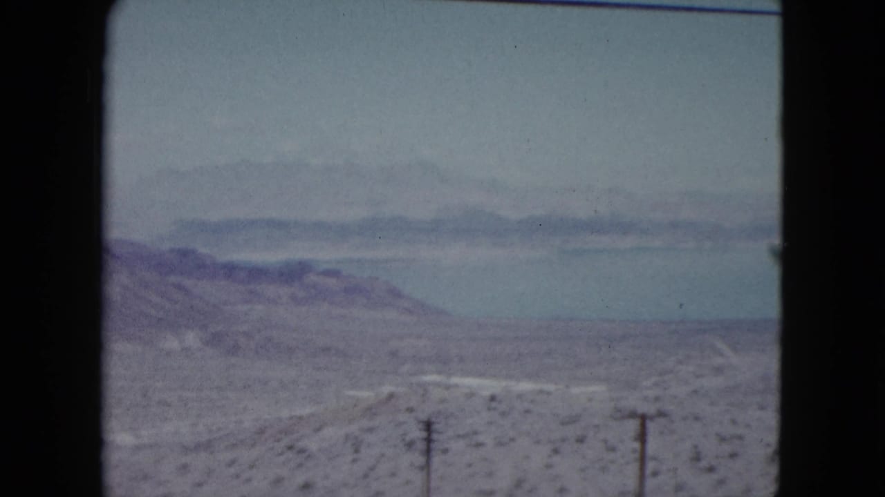1959 Nevada 8mm view across dry desert terrain toward a calm lake, low mountains, utility poles, and a pale winding road.