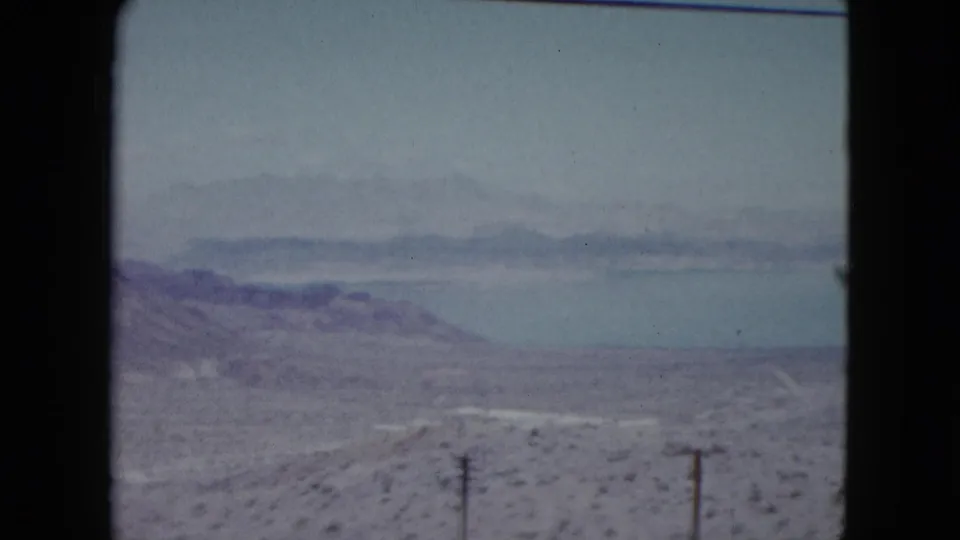1959 Nevada 8mm view across dry desert terrain toward a calm lake, low mountains, utility poles, and a pale winding road.