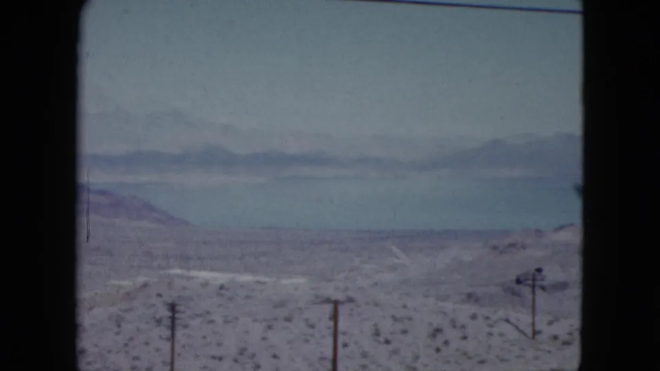 1959 Nevada home-movie frame with a broad lake, distant mountain ridge, dry desert foreground, sparse plants, and utility poles.