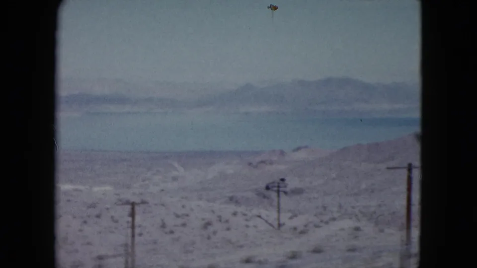 1959 Nevada 8mm frame showing dry scrubland descending toward a blue lake, with utility poles and distant hazy mountains.