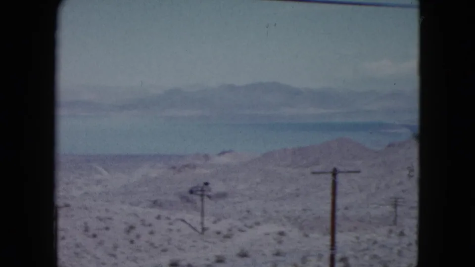 1959 Nevada 8mm view of arid hills, sparse brush, utility poles, and distant mountains seen from a moving vehicle.