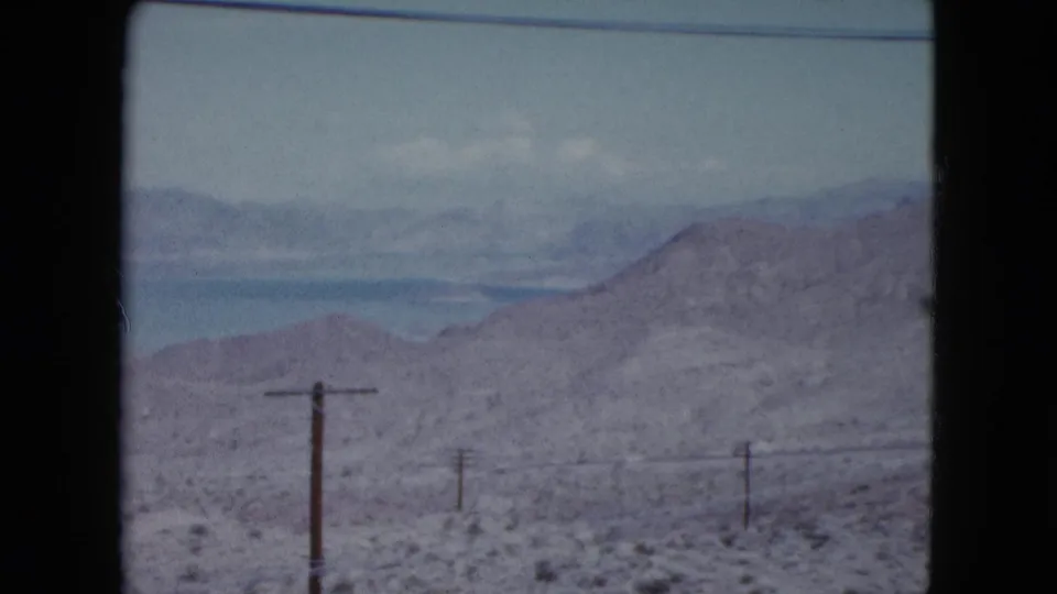 1959 Nevada roadside view with a nearer hillside, sparse brush, and wooden poles passing through the frame.