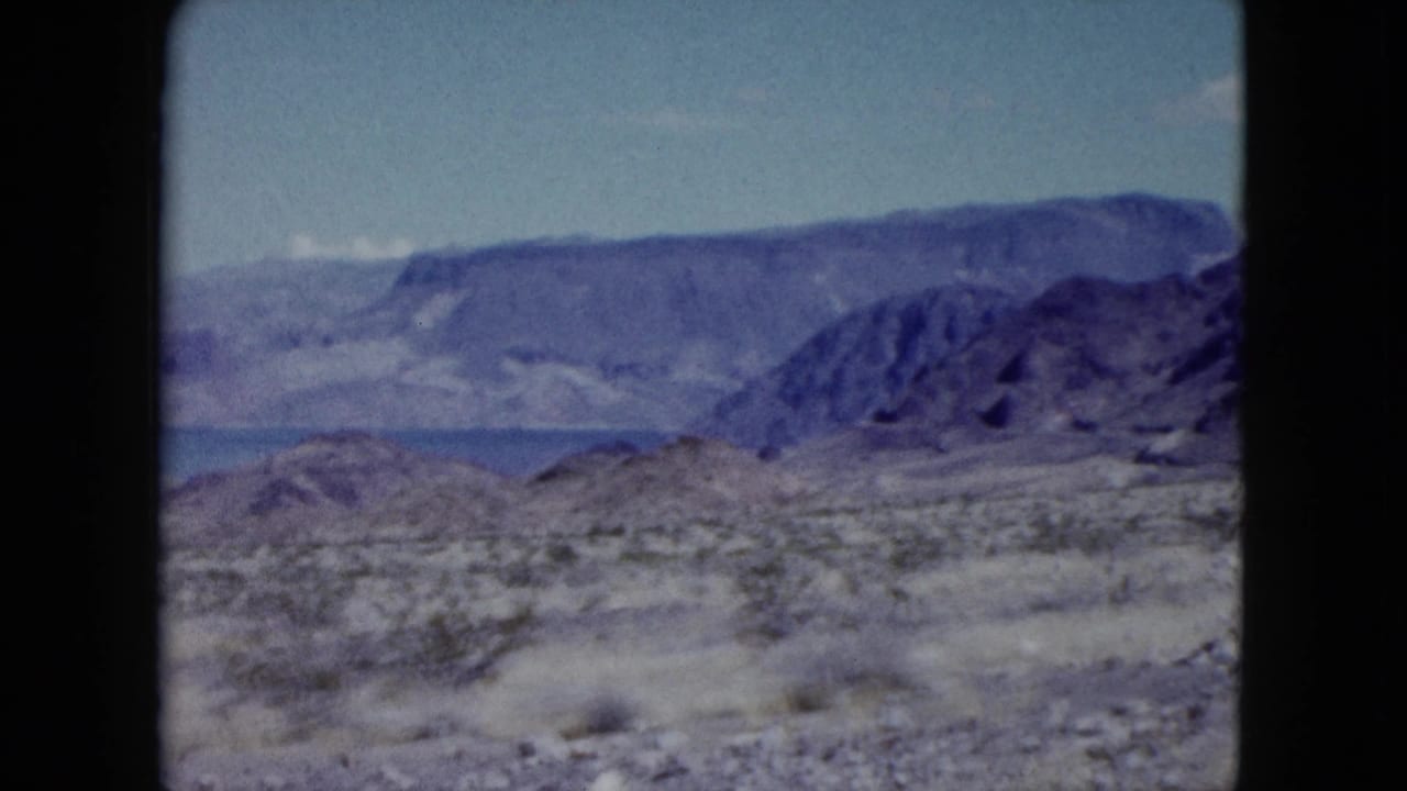 1959 Nevada 8mm frame showing arid scrubland, rocky terrain, a blue lake, and layered mountain ridges under pale sky.