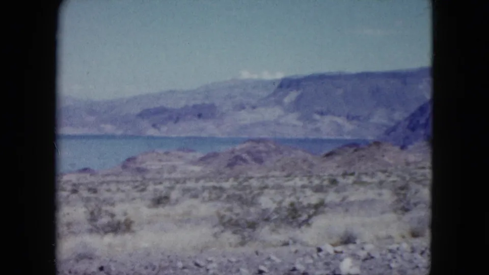 1959 Nevada home-movie frame with dry scrub terrain, blue lake water, and rugged mountain ridges in soft 8mm color.