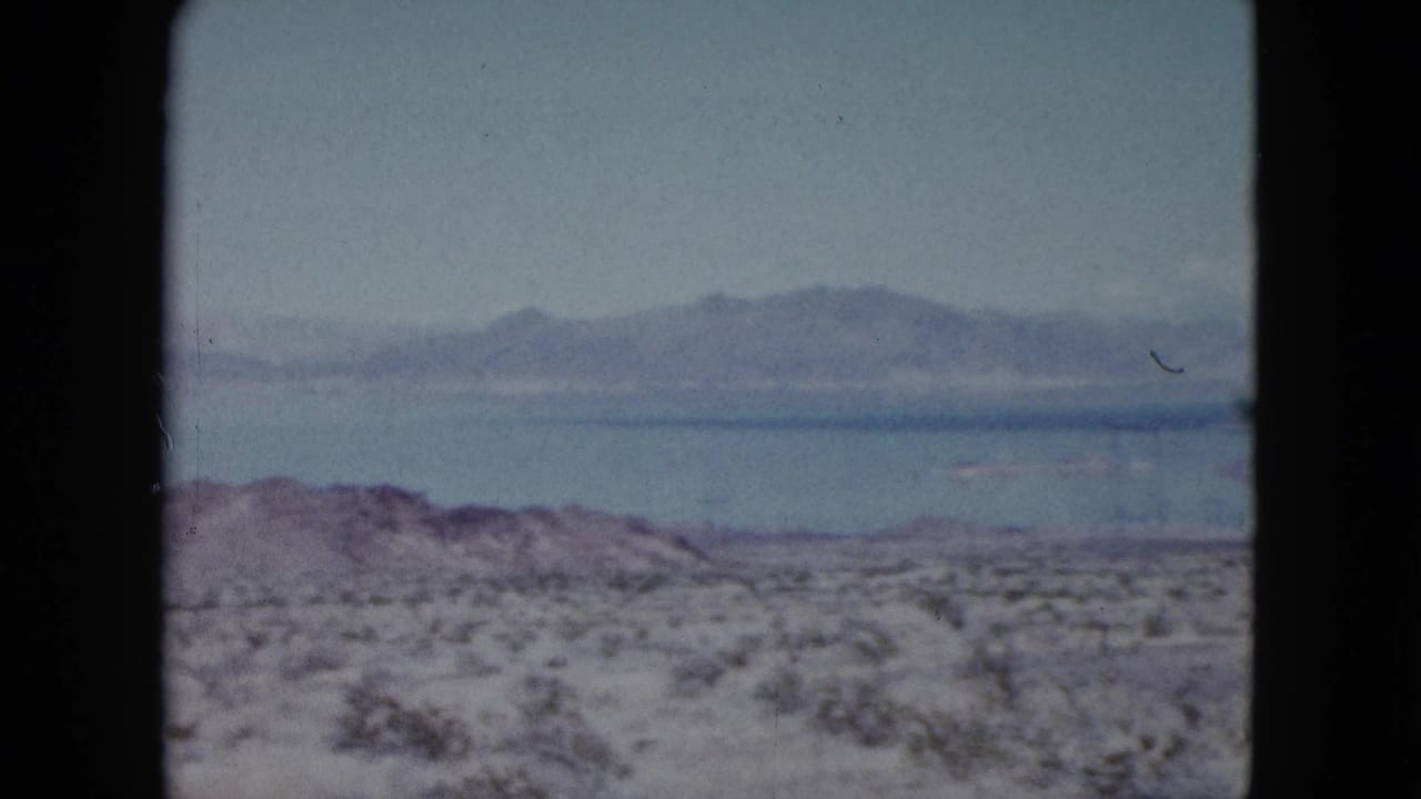 1959 Nevada 8mm frame showing scrub-covered desert foreground, a broad lake, and distant mountain ridges.
