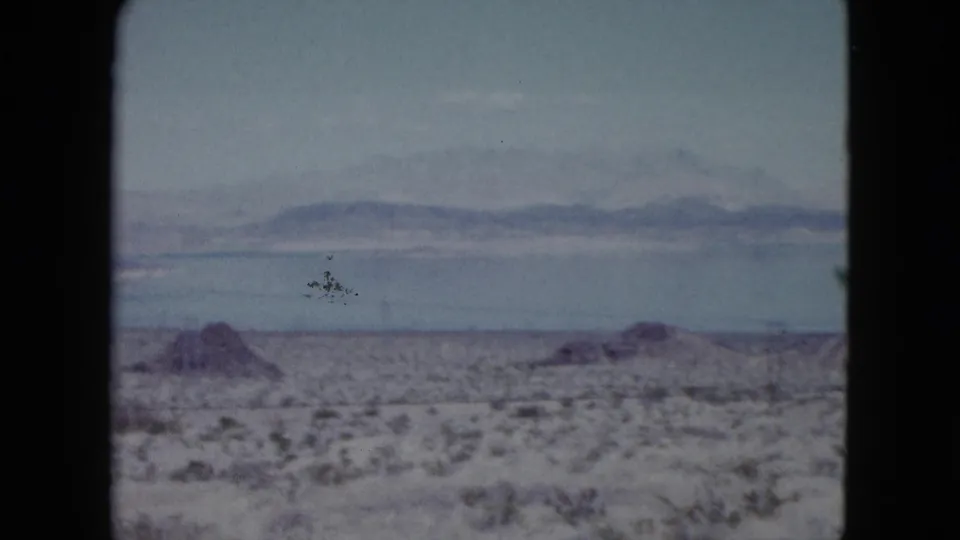 1959 Nevada home-movie frame with arid brushland, a narrow lake band, and hazy mountains beyond.