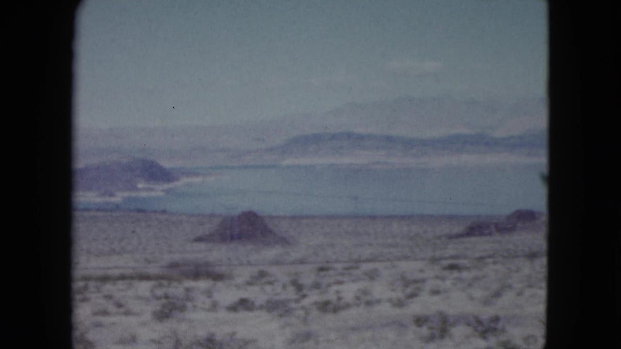 1959 Nevada 8mm frame showing desert scrub, rocky ground, distant blue water, and low mountains beyond the horizon.