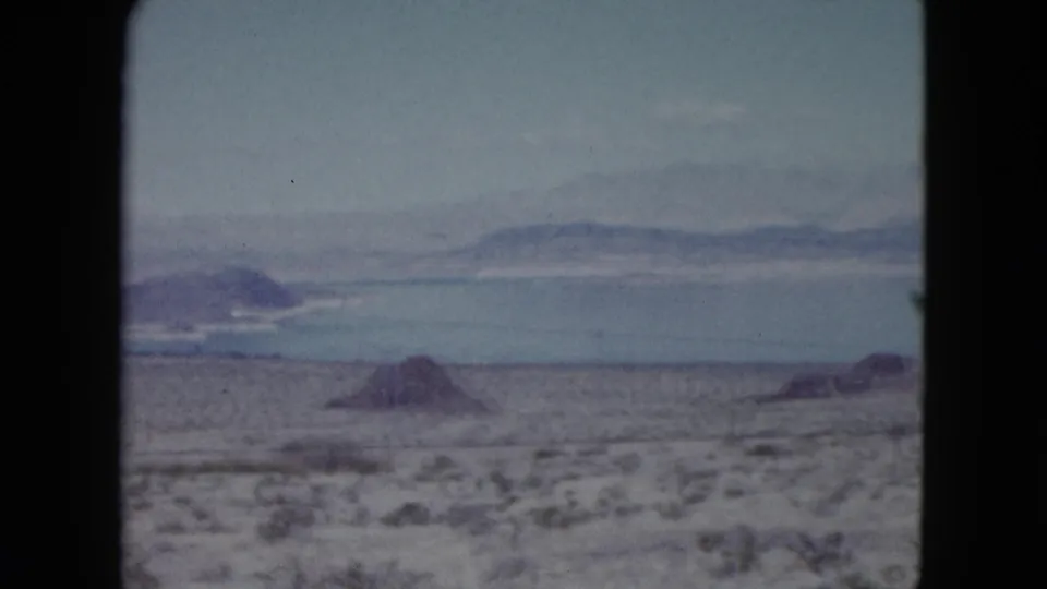 1959 Nevada 8mm frame showing desert scrub, rocky ground, distant blue water, and low mountains beyond the horizon.