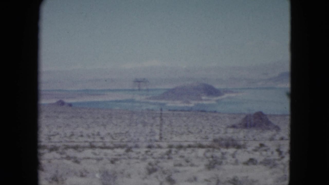 Nevada desert view with sparse vegetation, distant blue water, low mountains, and utility poles or towers visible in the distance.